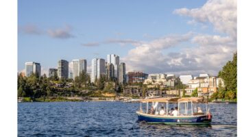 A small electric boat with people onboard cruises on Lake Washington in front of Bellevue, WA's modern skyline under a partly cloudy sky.
