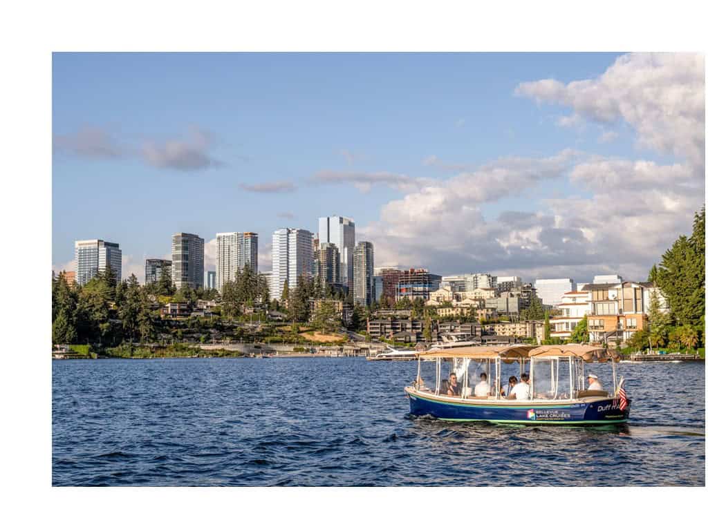 A small electric boat with people onboard cruises on Lake Washington in front of Bellevue, WA's modern skyline under a partly cloudy sky.