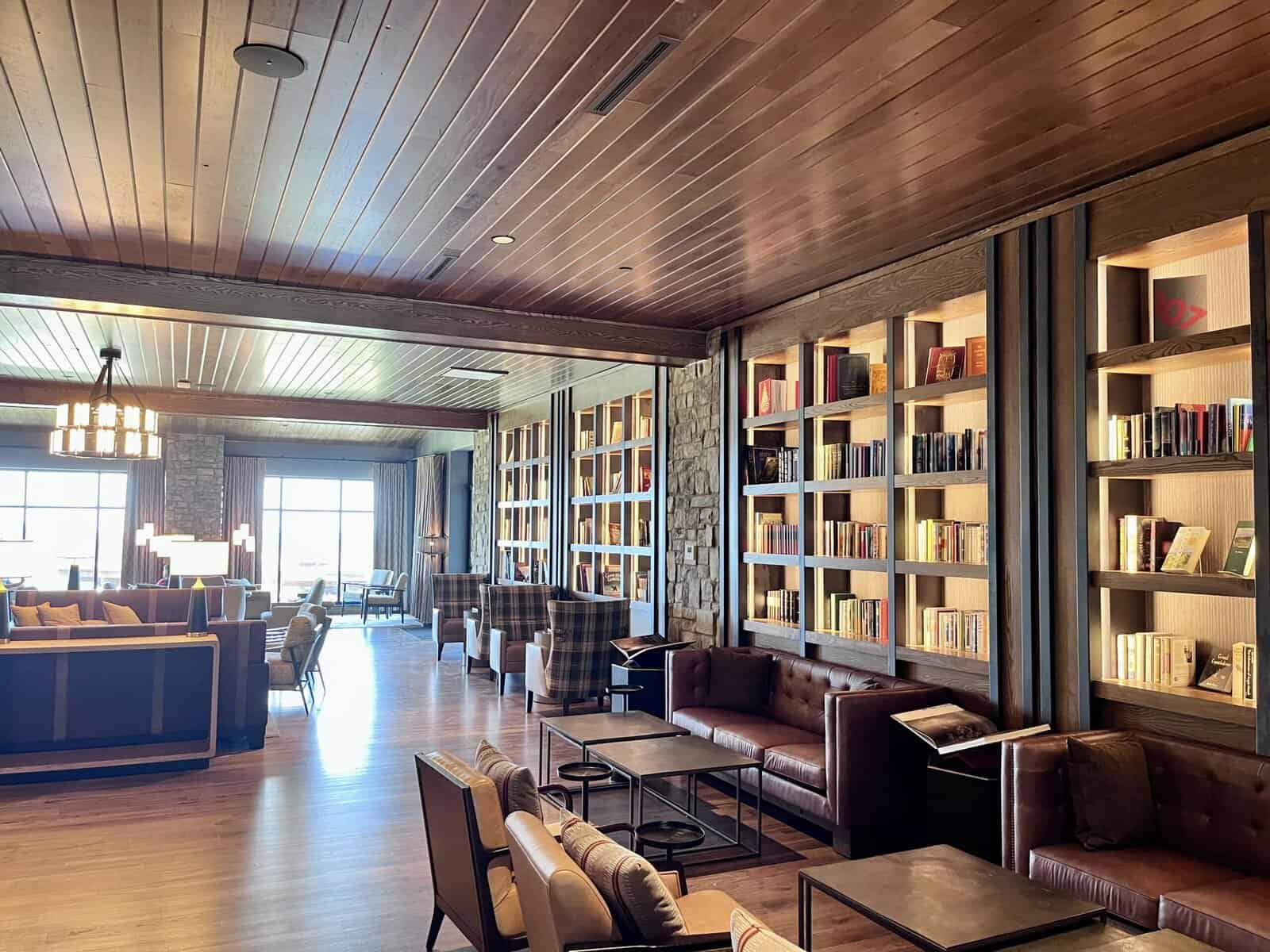 A wide shot of a cozy hotel library. On the right, tall, dark wood built-in bookshelves filled with books are illuminated by integrated lighting. In front of the bookshelves are several brown leather sofas and coffee tables. To the left, more seating areas with armchairs and couches are visible, and in the background, large windows offer a view of mountains and sky. The ceiling is made of warm-toned wood planks, and the floor is also wood.