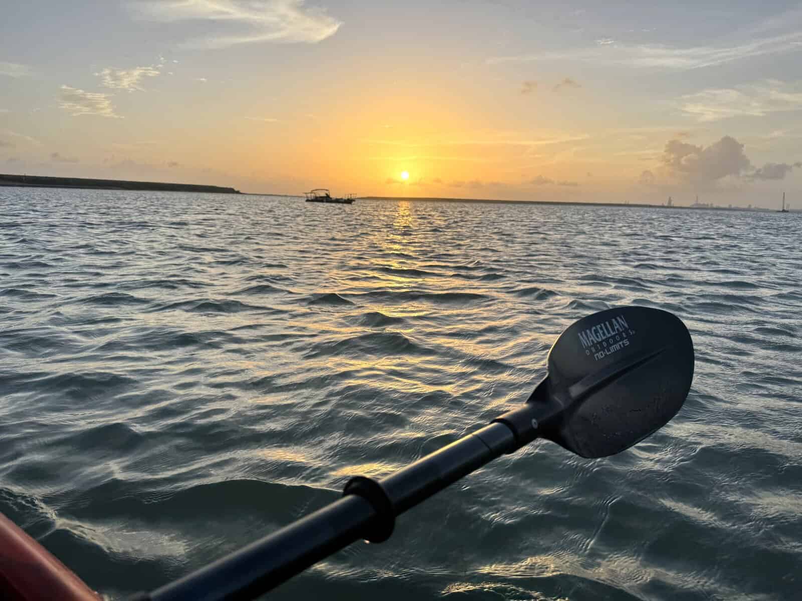 A kayak paddle rests atop the water as the sun sets along the horizon.
