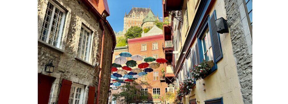 Colorful umbrellas hang above Rue du Petit-Champlain with Château Frontenac rising in the background.