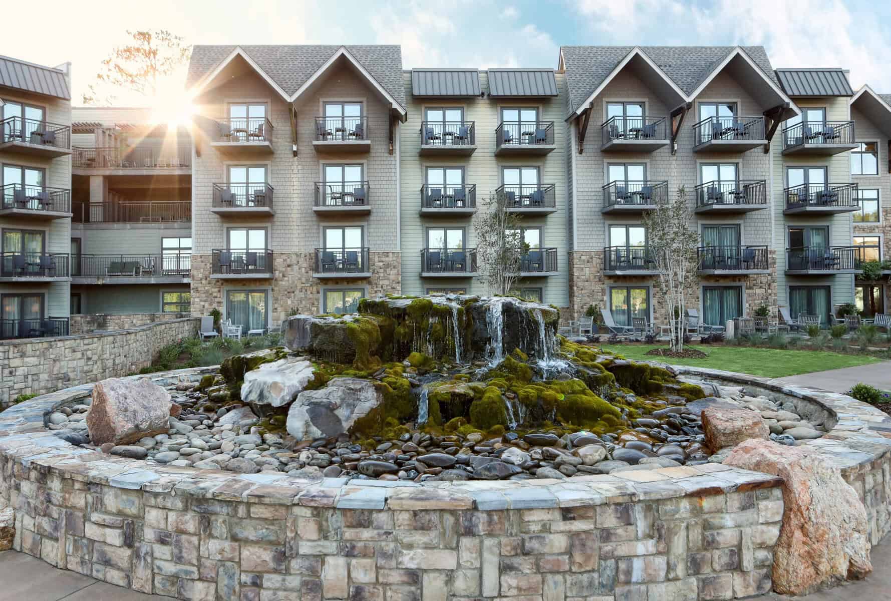 Exterior view of a multi-story lodge at Callaway Gardens with balconies overlooking a landscaped courtyard, highlighted by a moss-covered stone fountain with cascading water and surrounded by trees and greenery.