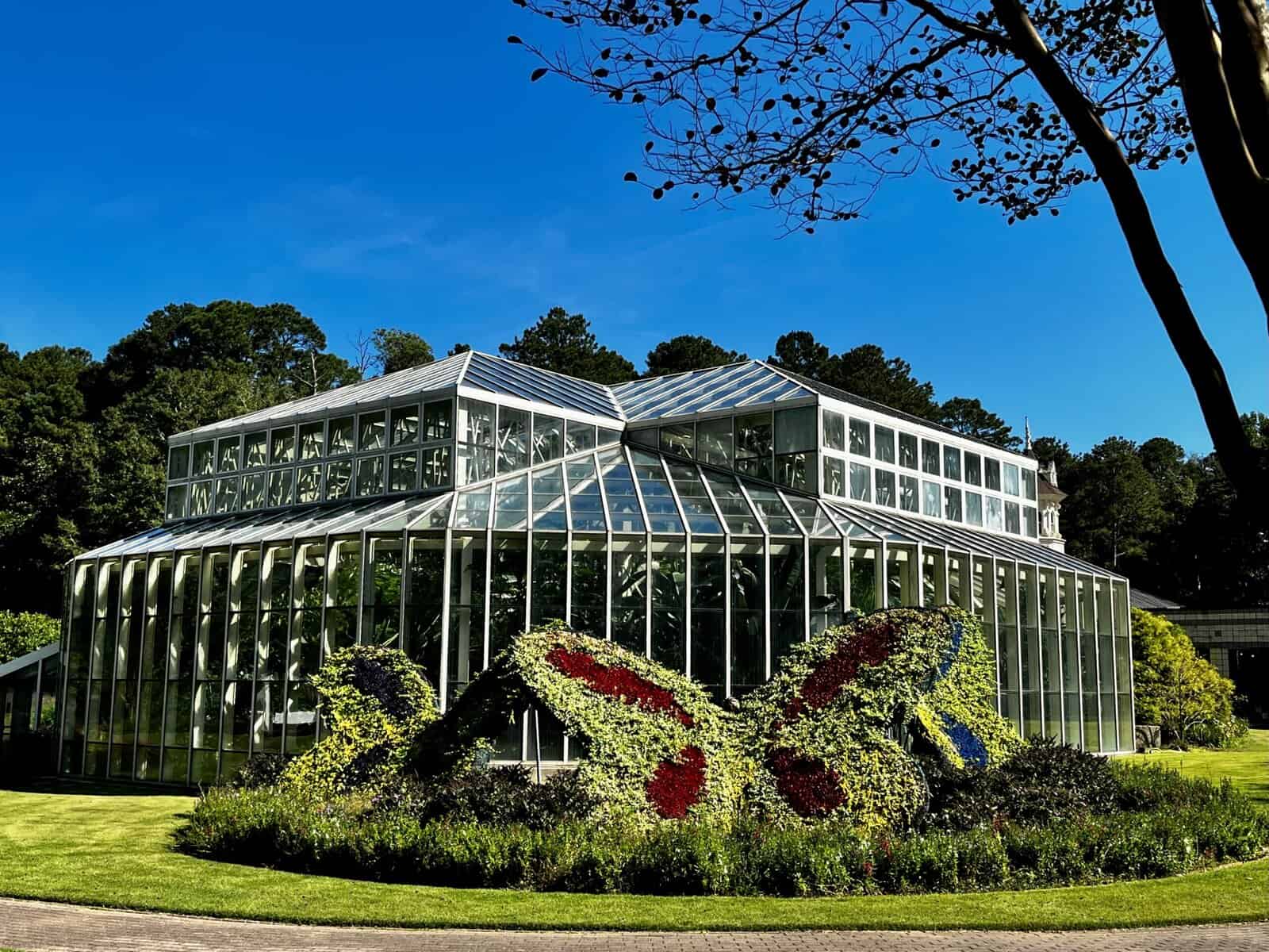 Bright, sunny exterior of the Cecil B. Day Butterfly Center at Callaway Resort & Gardens, showcasing a striking glass conservatory surrounded by lush gardens and large, colorful butterfly-shaped topiaries, all set against a backdrop of towering green trees and a clear blue sky.