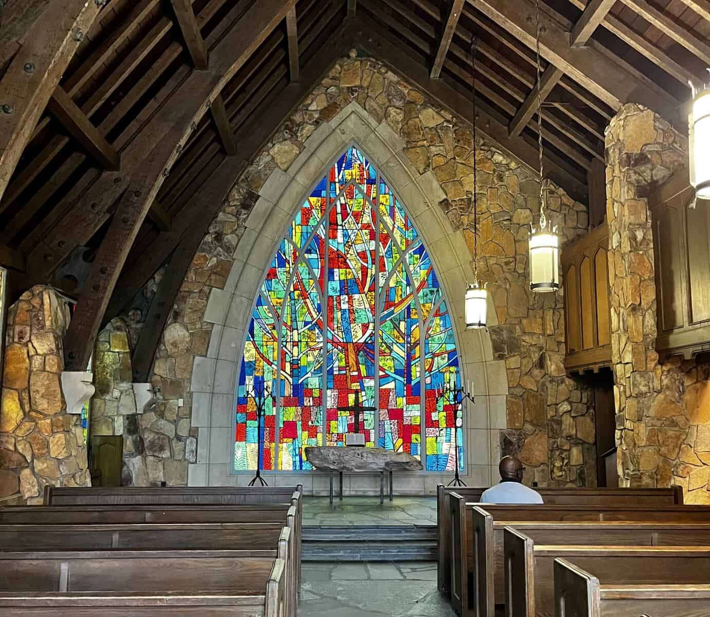Interior view of a rustic stone chapel with exposed wooden beam ceiling, featuring a large colorful stained glass window in an abstract tree pattern as the focal point behind the altar, with wooden pews arranged in rows and warm lighting throughout the space.
