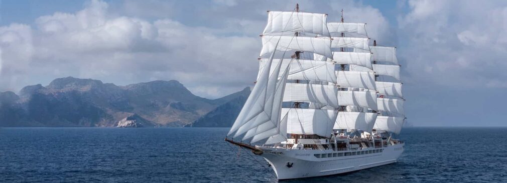A large white sailing ship with all sails unfurled sailing near a mountainous coastline.