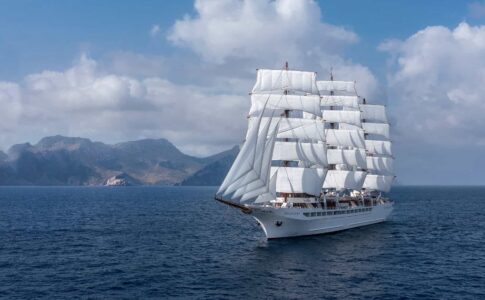 A large white sailing ship with all sails unfurled sailing near a mountainous coastline.