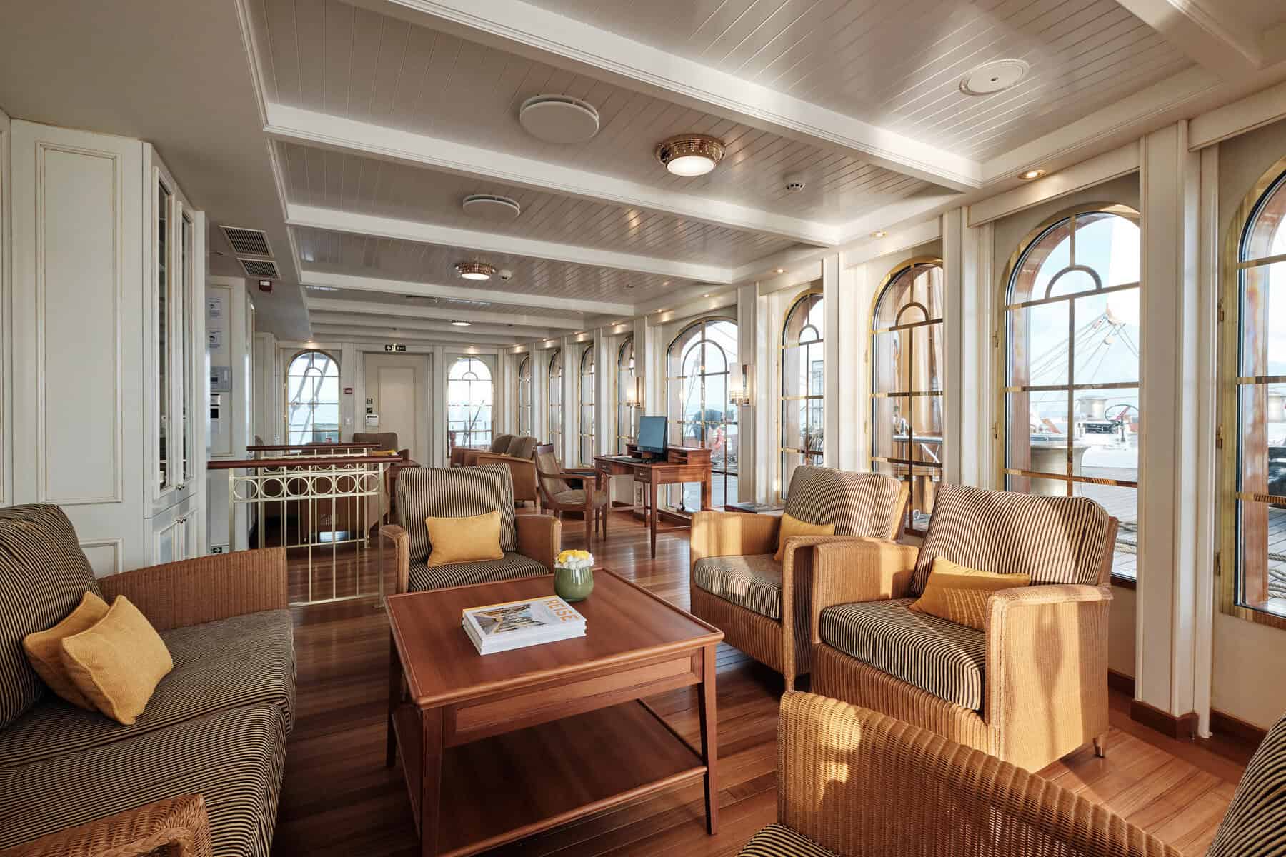 A luxury ship lounge with striped furniture, wooden floors, and large arched windows overlooking the ocean.