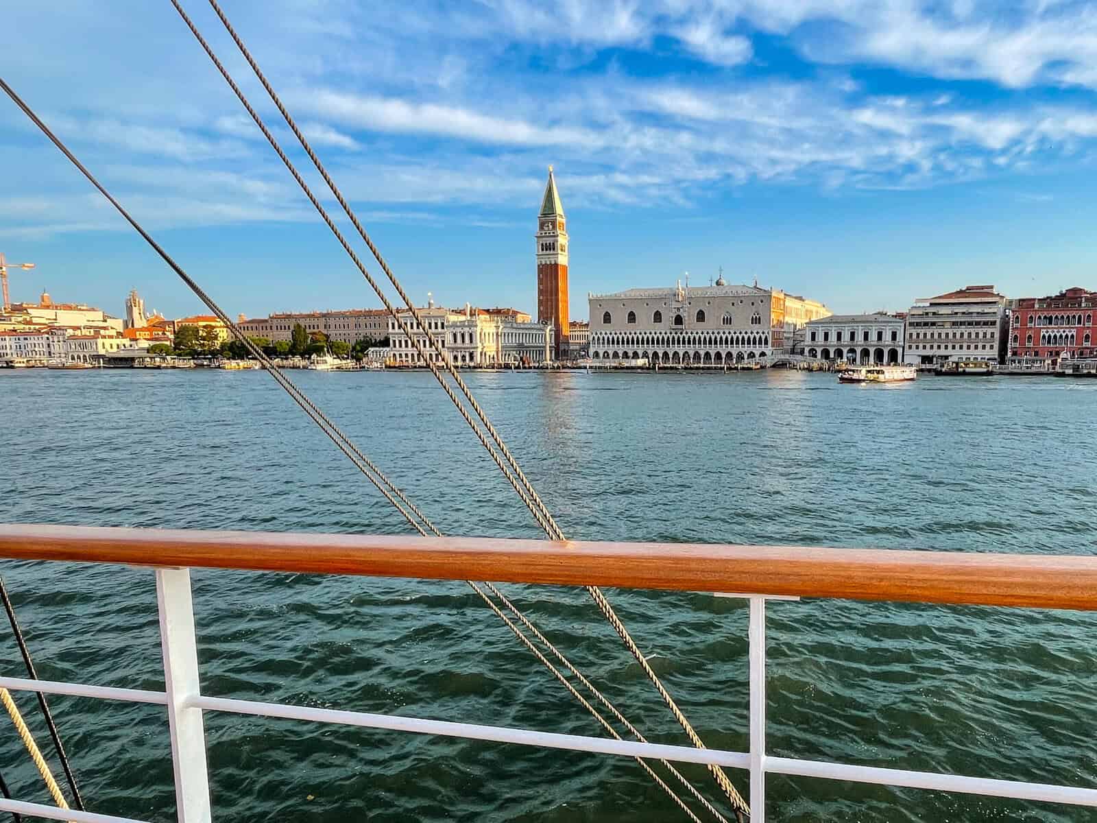 View of Venice waterfront and bell tower from a sailing ship deck with rigging visible in foreground.