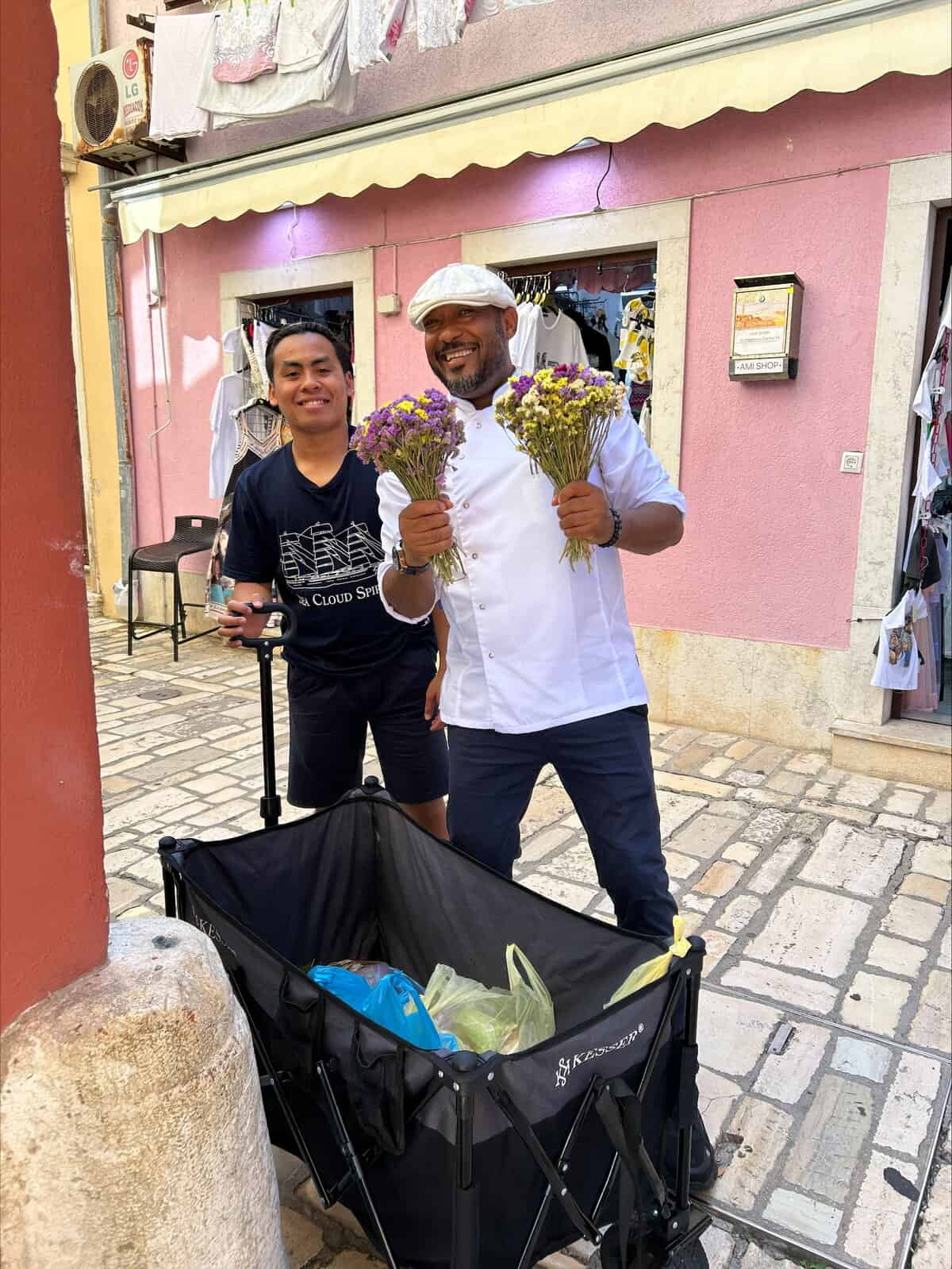 Two chefs in white uniforms holding bouquets of purple and yellow flowers, standing with a black rolling cart on a cobblestone street.