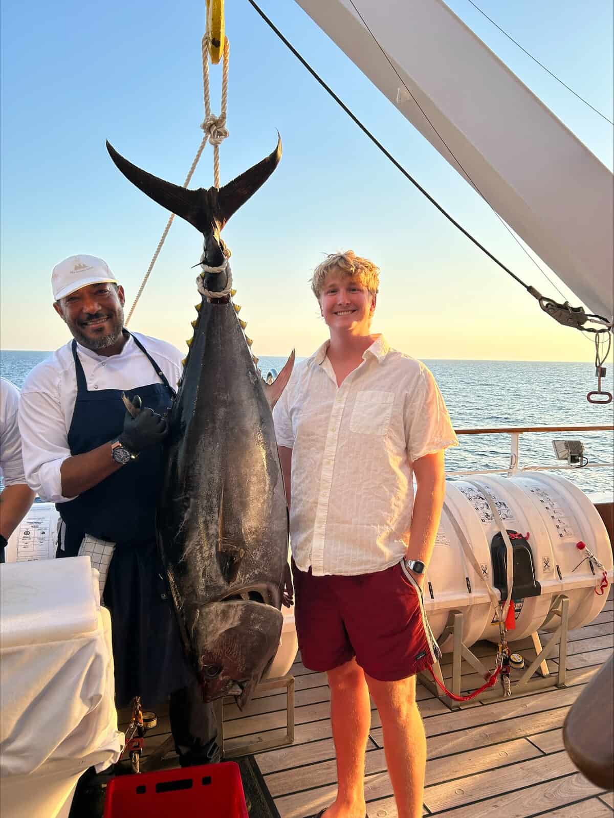 Chef and guest standing on ship deck at sunset with a large tuna hanging between them.