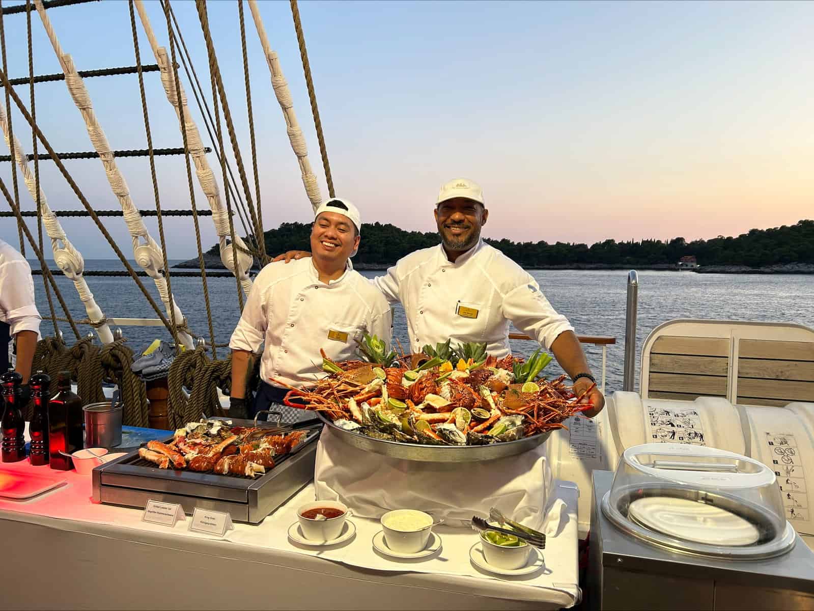 Two chefs in white uniforms presenting large platters of grilled seafood and shellfish on the ship's deck at dusk