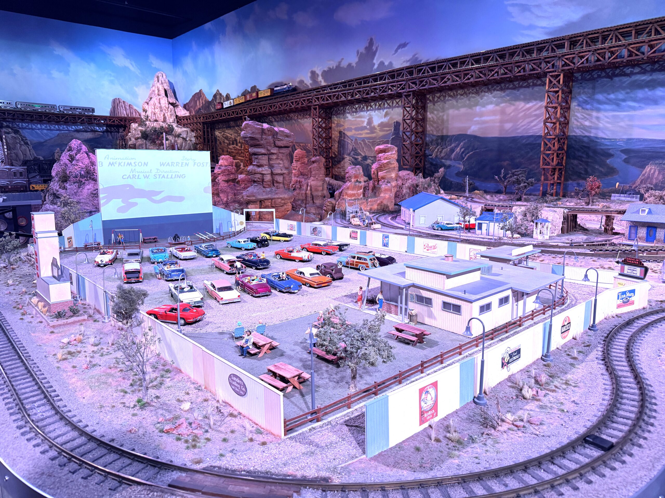 A model railroad track snakes around a drive-in movie theater complete with cars, benches, and customers.