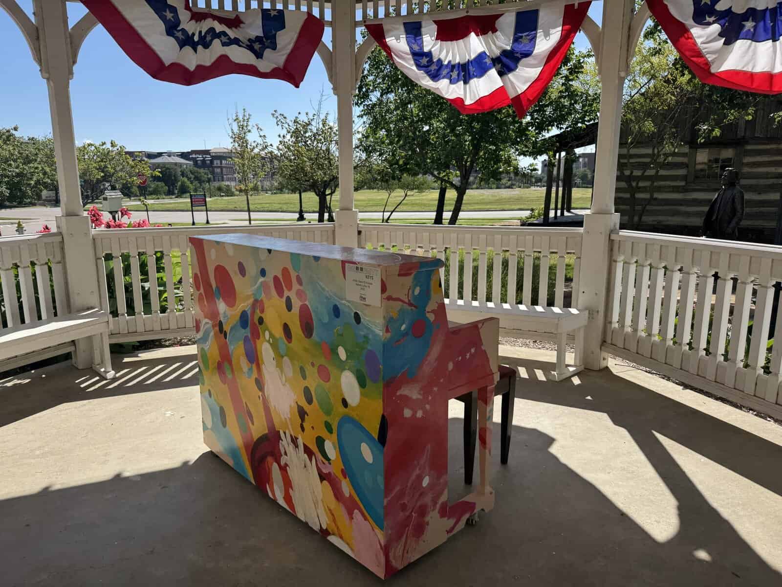 A colorful piano sitting underneath a white gazebo. A log cabin stands in the background.