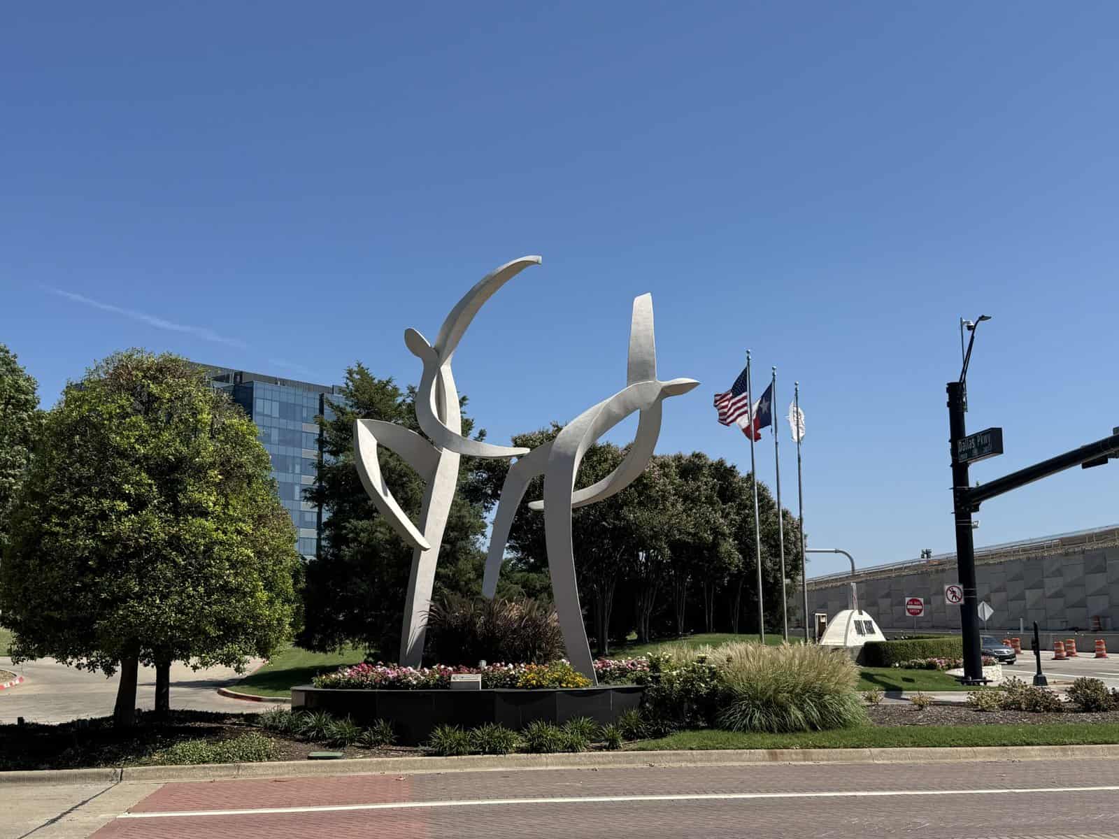 Two large white roadside sculptures shaped like dancers, surrounded by flags and trees.