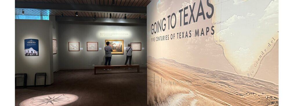 Interior gallery at the Briscoe Western Art Museum showing the exhibition title wall for “Going to Texas: Five Centuries of Texas Maps,” framed maps on the far wall, two visitors viewing the works, and a compass graphic projected on the floor.