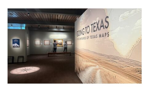 Interior gallery at the Briscoe Western Art Museum showing the exhibition title wall for “Going to Texas: Five Centuries of Texas Maps,” framed maps on the far wall, two visitors viewing the works, and a compass graphic projected on the floor.