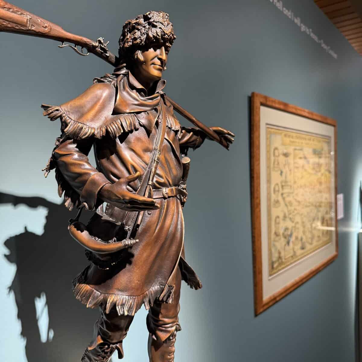 Close view of a bronze statue of Davy Crockett—wearing a coonskin cap and carrying a rifle over his shoulder—with a framed Texas Centennial map hanging on the teal gallery wall behind him.