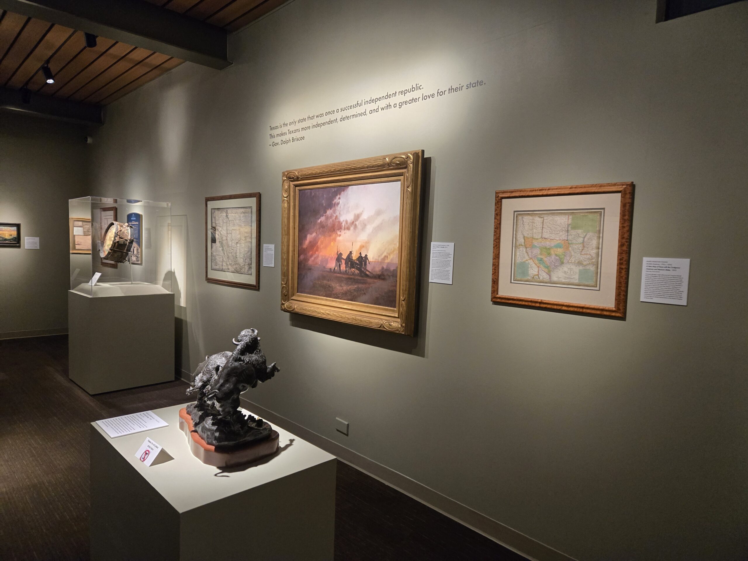 Wide gallery view of the Briscoe’s “Going to Texas” exhibition showing a foreground bronze of a cowboy on a bucking horse, a central large oil painting of riders at sunset in a gold frame, framed historic Texas maps on either side with wall labels, and a glass case to the left holding a drum and documents; above, wall text features a quote by Gov. Dolph Briscoe.