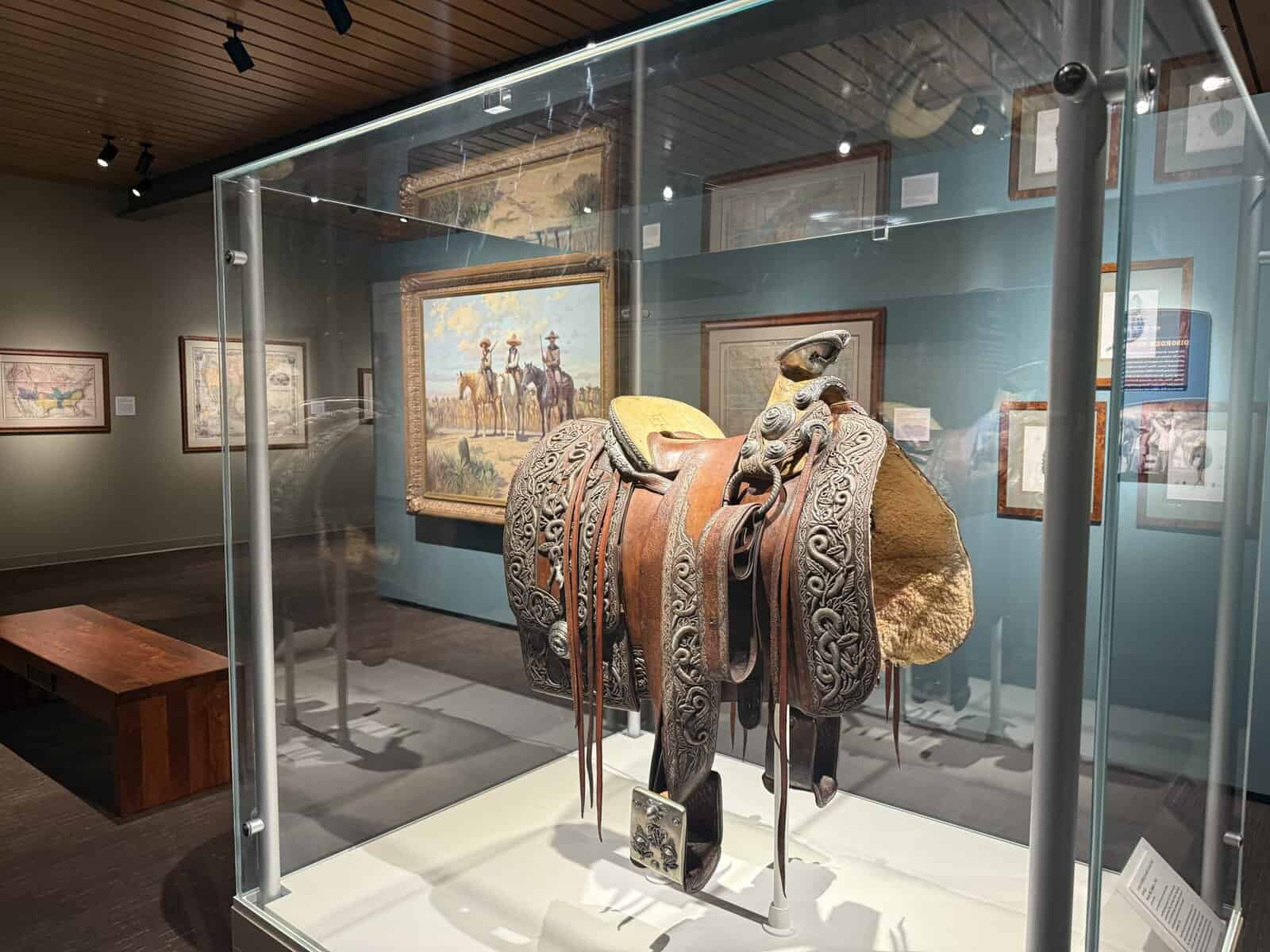 Glass case displaying Pancho Villa’s richly tooled leather saddle with silver accents; surrounding gallery walls feature framed Texas maps and Western paintings at the Briscoe Western Art Museum.