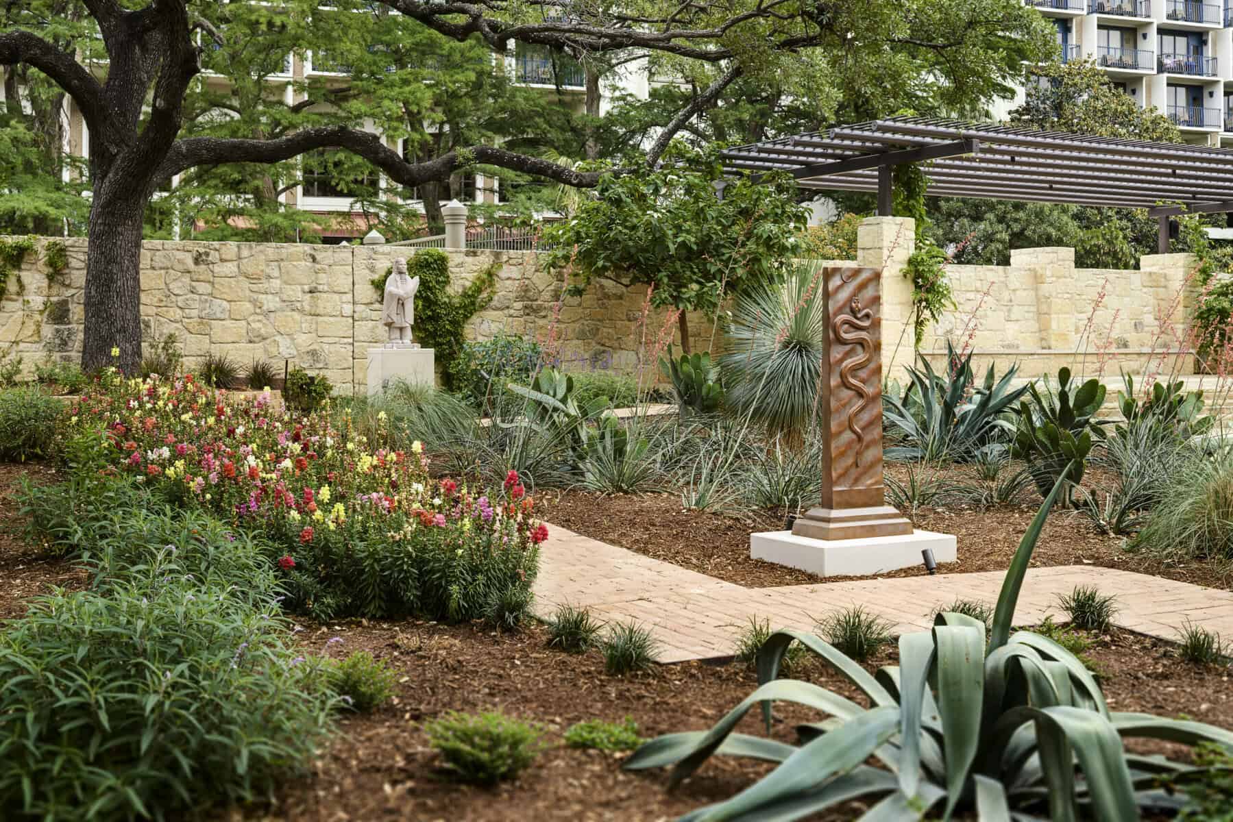 Outdoor view of the Briscoe’s McNutt Sculpture Garden featuring flowering beds, agave and yucca, a bronze column with a snake motif, a figurative statue in the distance, limestone walls, and a shaded pergola beneath spreading live oaks.