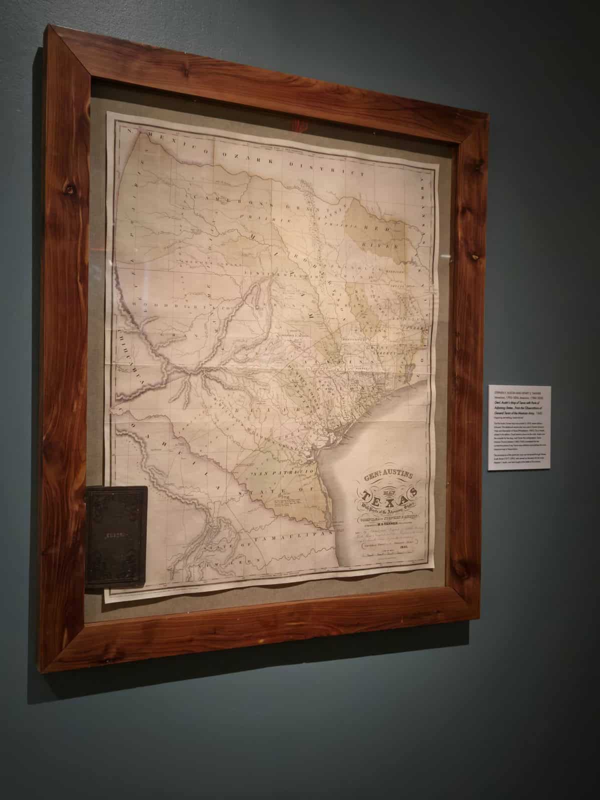 Framed historical pocket map titled “Genl. Austin’s Map of Texas,” shown fully opened with visible fold lines; a small black pocket case is mounted at the lower left inside the frame, with a wall label beside the display.