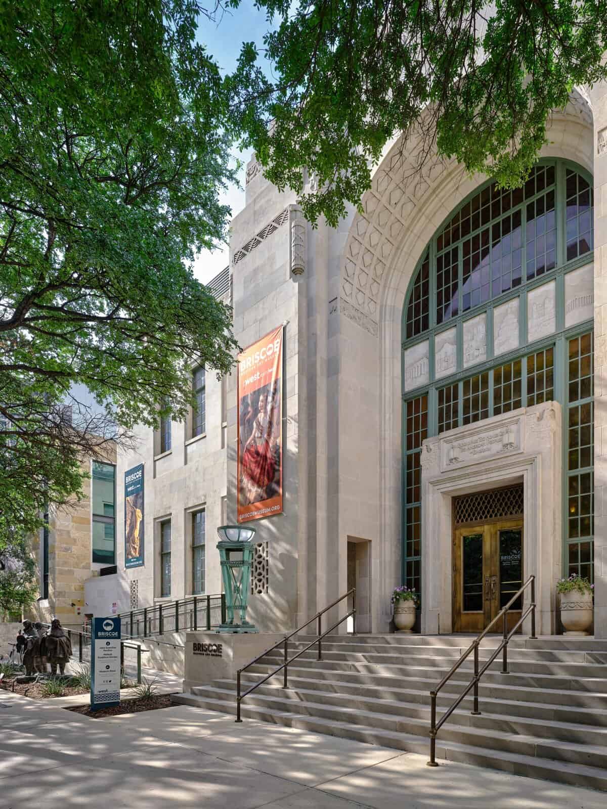 Exterior view of the Briscoe Western Art Museum showing its limestone façade, arched main entrance, steps and trees in front.