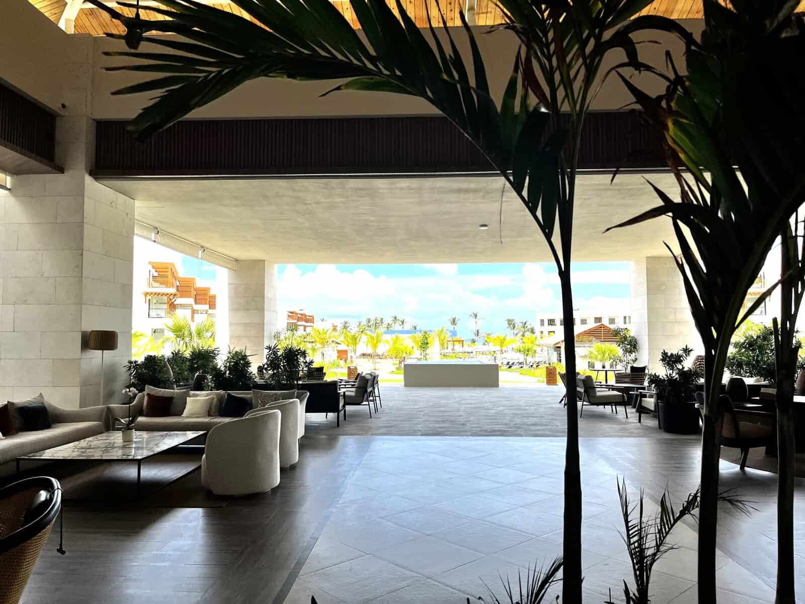 Modern open-air resort lobby with high ceilings, neutral stone columns, contemporary seating areas with plush couches and chairs, potted palm plants, and ocean views visible through the wide opening leading to the pool and beach area beyond.