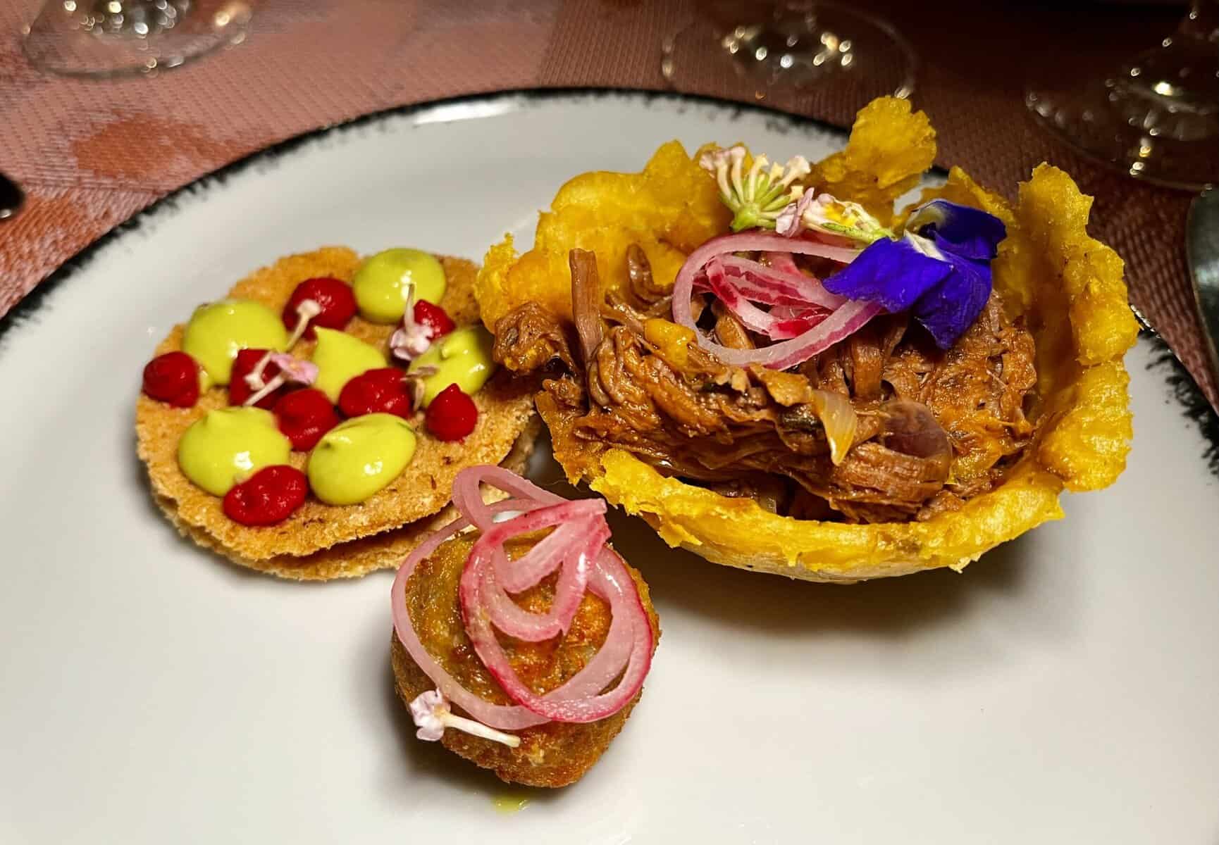 Plated Dominican appetizers featuring a croquette, a fried plantain cup filled with braised beef topped with pickled onions and edible flowers, and cassava bread rounds garnished with avocado mousse dots and microgreens on a white plate with dark rim.