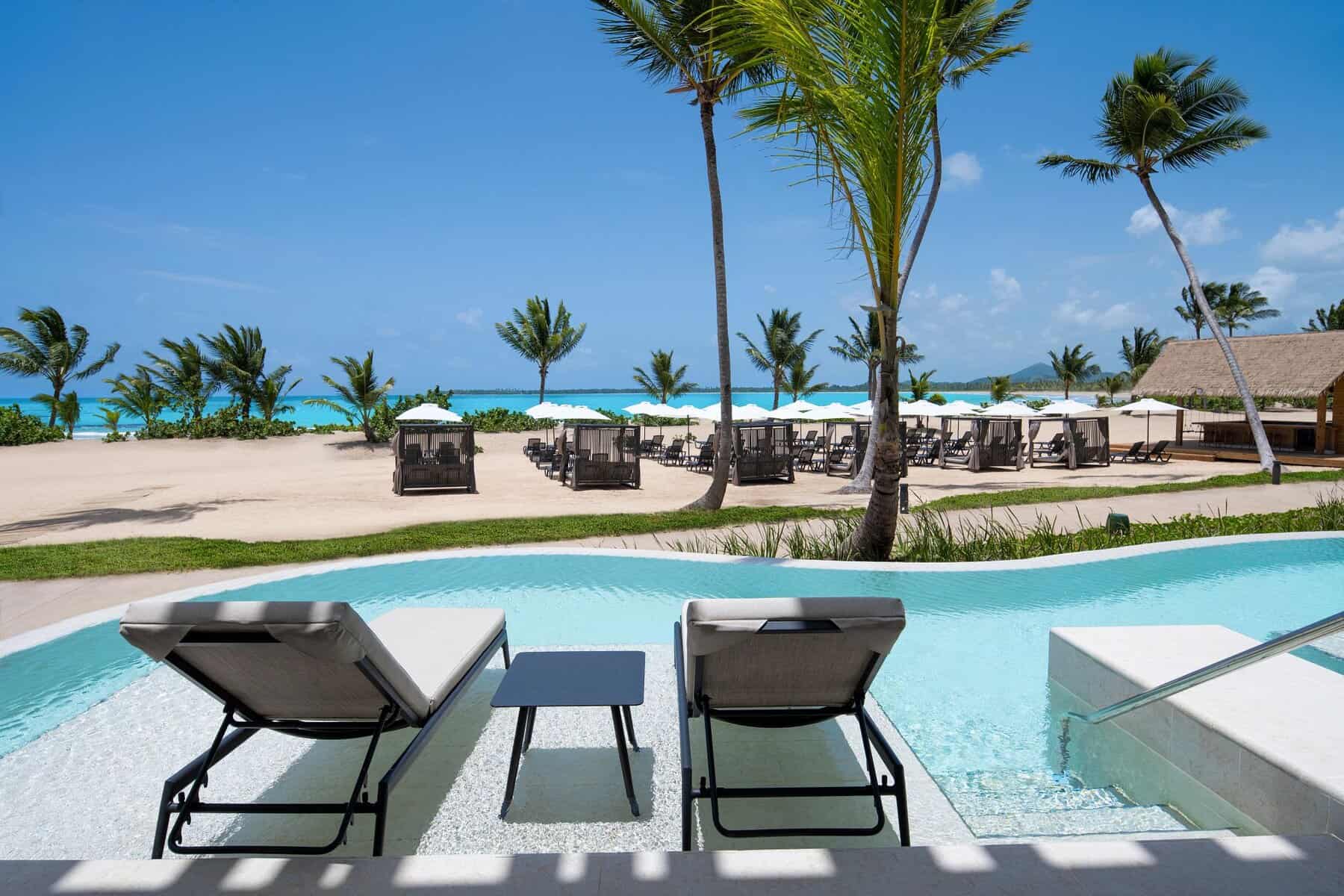 Two modern pool loungers with white cushions on a submerged platform in a private swim-out suite section of the pool, overlooking pristine white sand beach with cabanas, palm trees, and bright blue ocean under clear sky at Secrets Playa Esmeralda Resort.