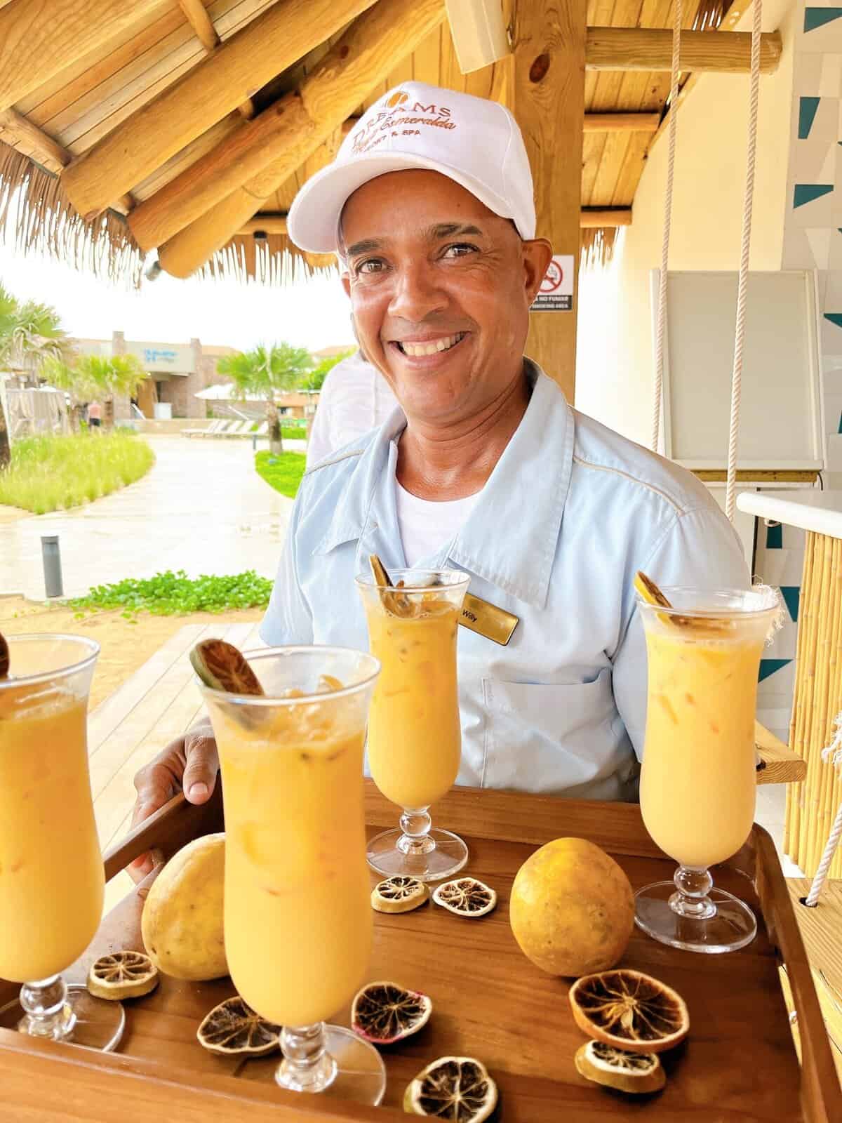 Smiling resort bartender in white cap and light blue shirt presents a wooden tray with four tropical yellow cocktails garnished with passion fruit and dried citrus slices at a thatched-roof beach bar.