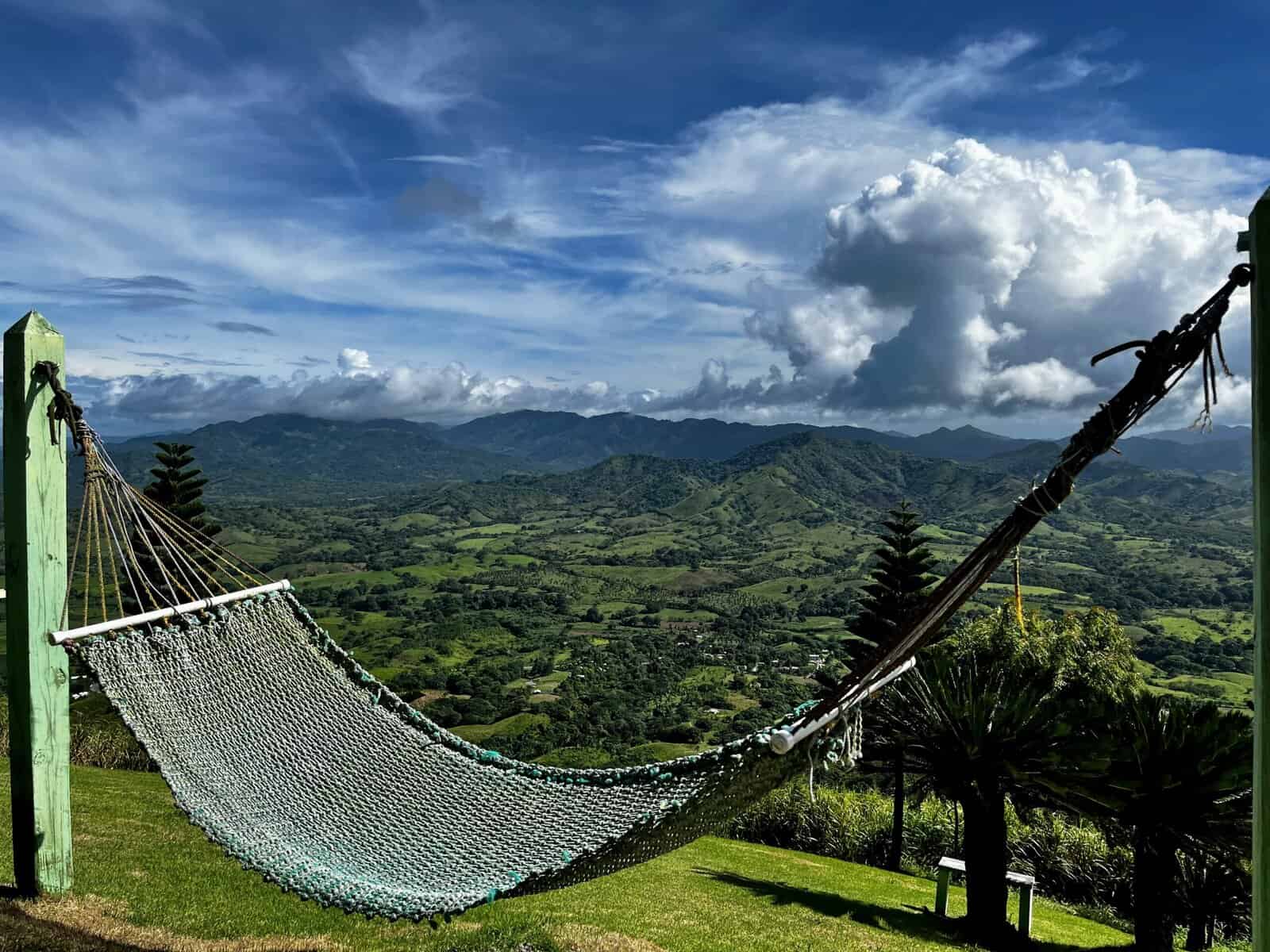 Woven hammock suspended between green wooden posts overlooking lush green mountains and valleys of the Dominican Republic countryside under a dramatic blue sky with billowing white clouds.