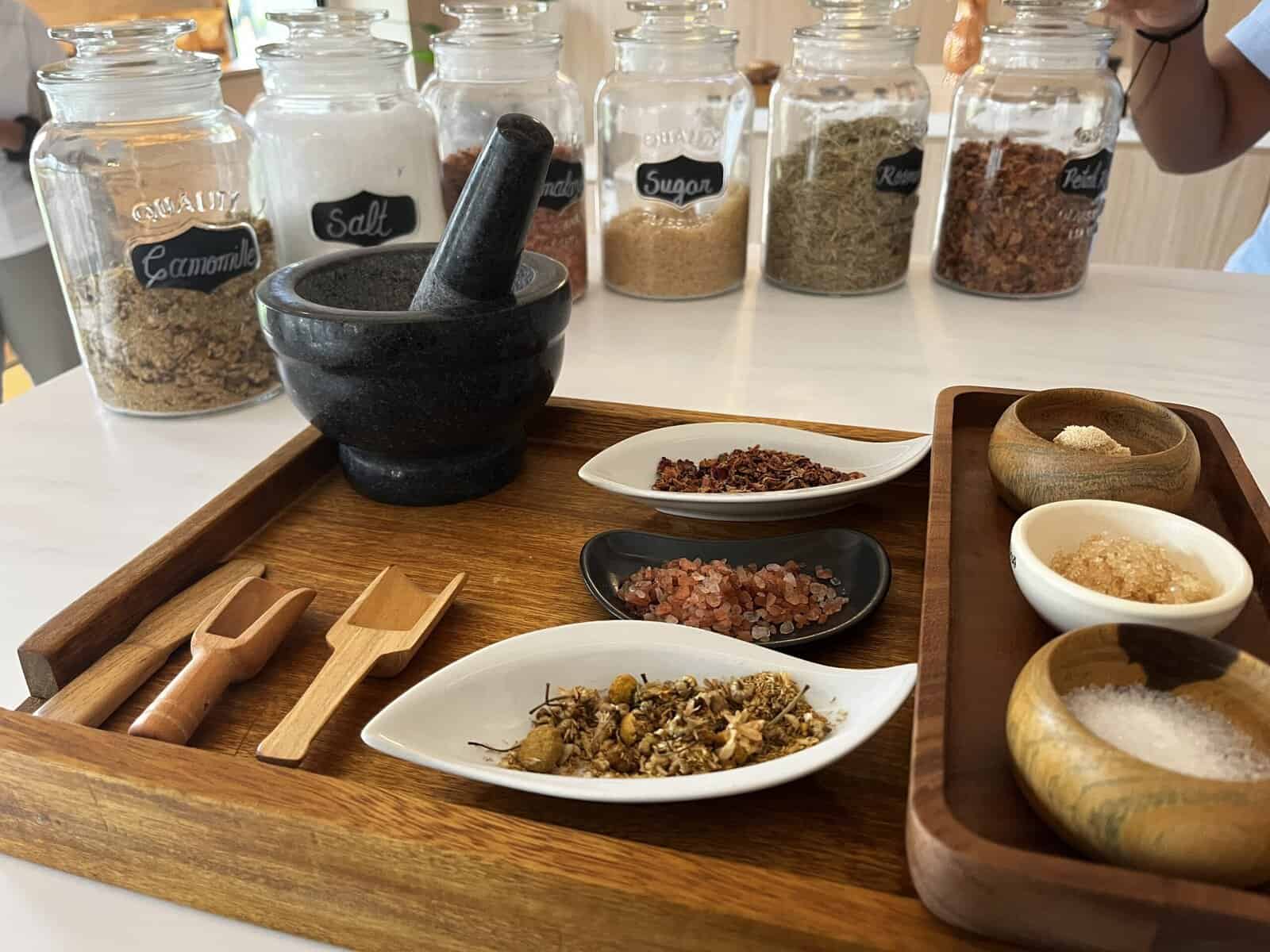 Spa body scrub making station with wooden tray displaying mortar and pestle, white dishes containing dried botanicals and pink salt crystals, wooden bowls with various scrub ingredients, and glass jars of herbs and spices including chamomile, salt, sugar, and rosemary in the background.
