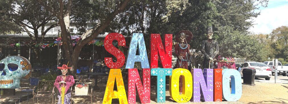 “San Antonio” spelled out in colorful block letters, adorned by catrin and catrina skeletons and a smiling skull with a butterfly design.