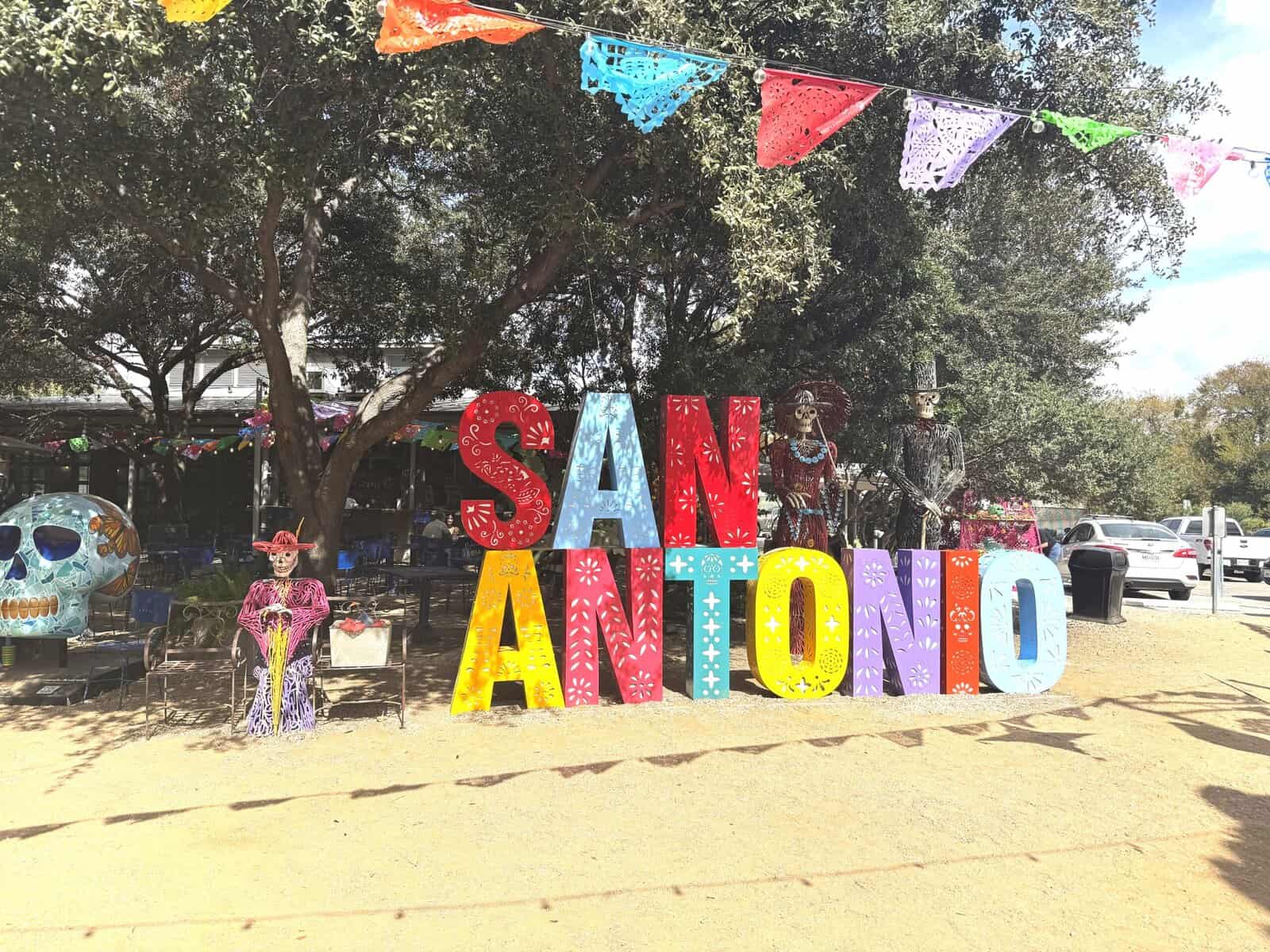 “San Antonio” spelled out in colorful block letters, adorned by catrin and catrina skeletons and a smiling skull with a butterfly design.