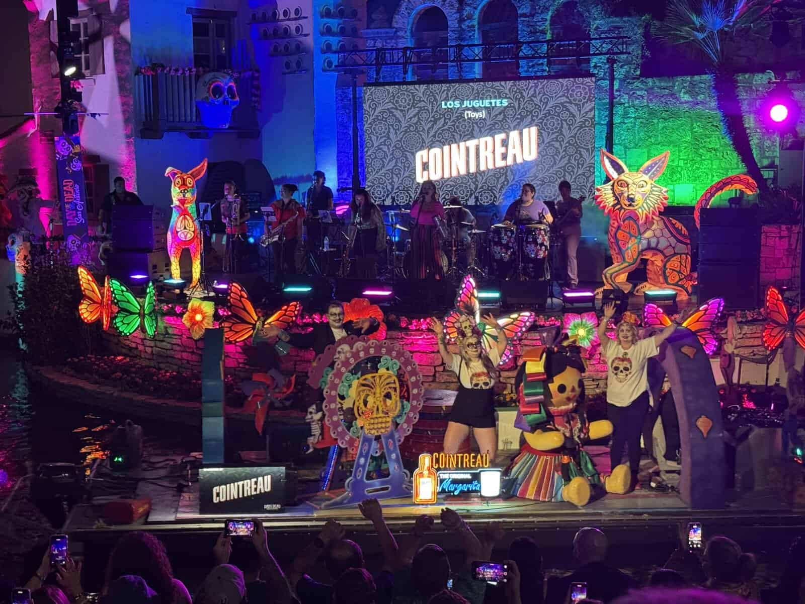 A parade float on the river featuring dancers and toys, with musicians performing on a stage in the background.