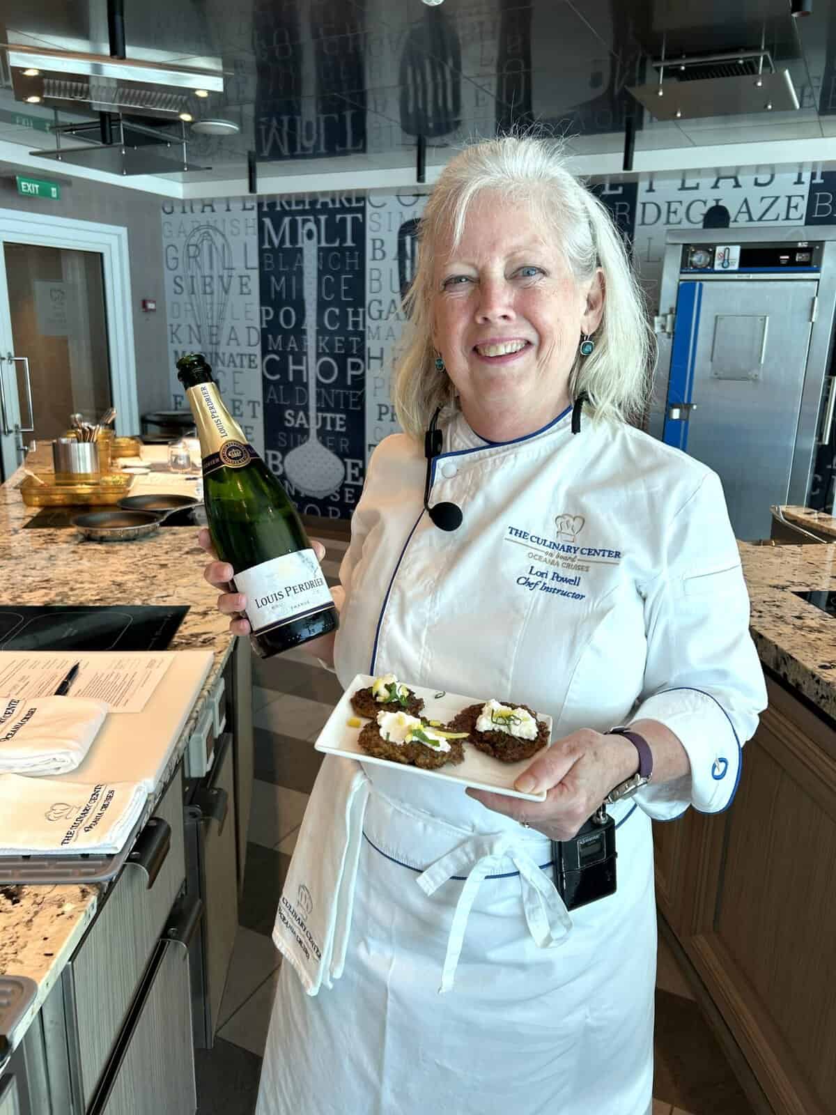 Chef instructor in white chef's coat holding champagne bottle and plate of appetizers in professional kitchen.