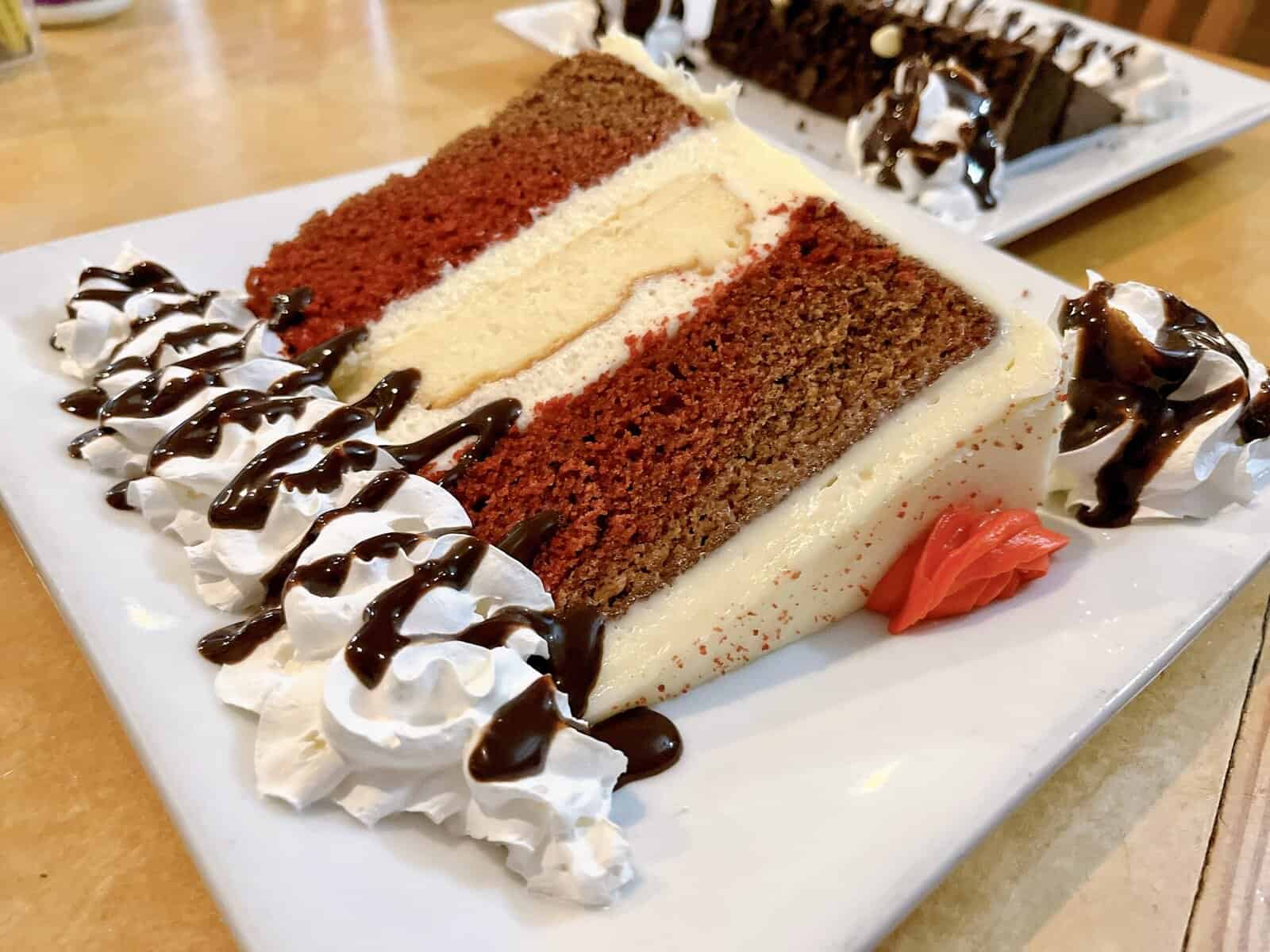 Close-up of a layered cake slice showing red velvet cake layers separated by thick white cheesecake and cream cheese frosting, garnished with whipped cream rosettes, chocolate drizzle, and a small decorative red frosting rose, with a chocolate cake slice visible in the background.