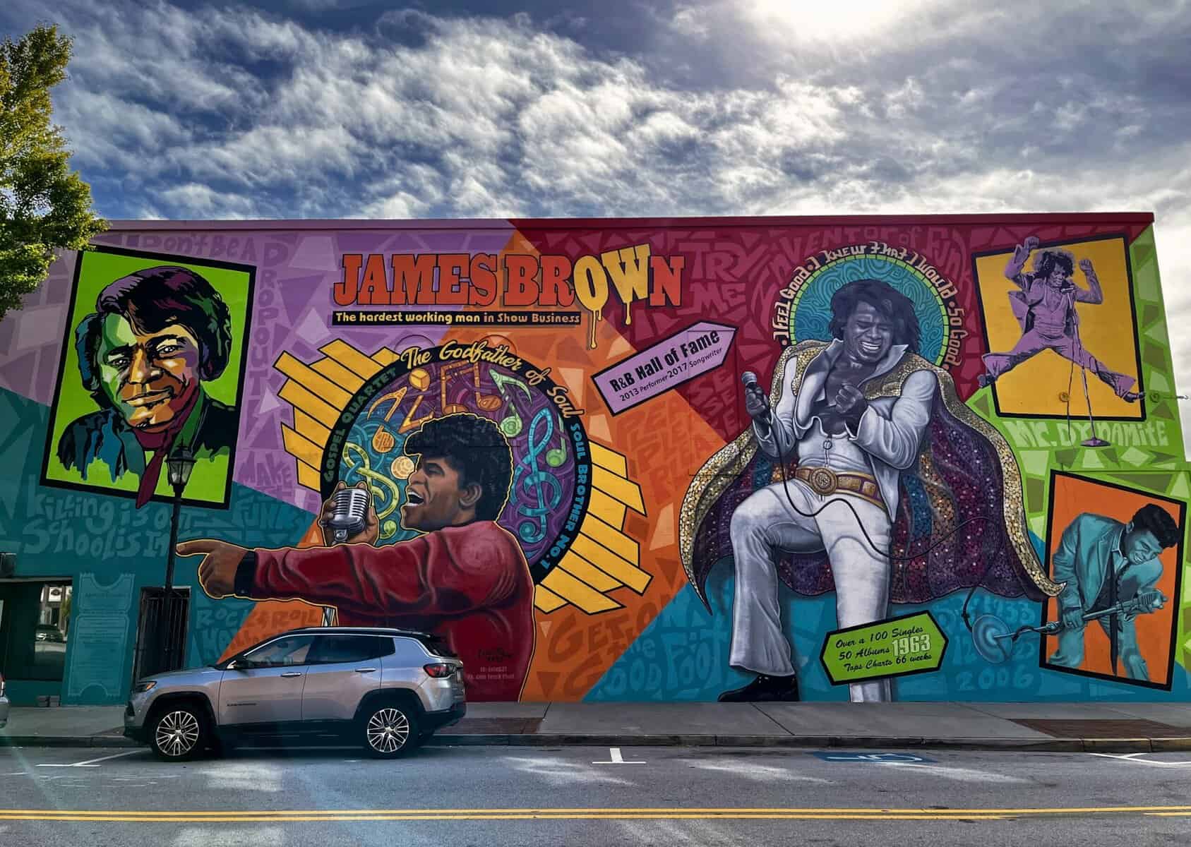 Colorful street mural celebrating James Brown featuring multiple portraits of the performer in vibrant shades of purple, orange, yellow, and green, with text reading "JAMES BROWN The hardest working man in Show Business" and "The Godfather of Soul," alongside references to his R&B Hall of Fame induction in 2013.