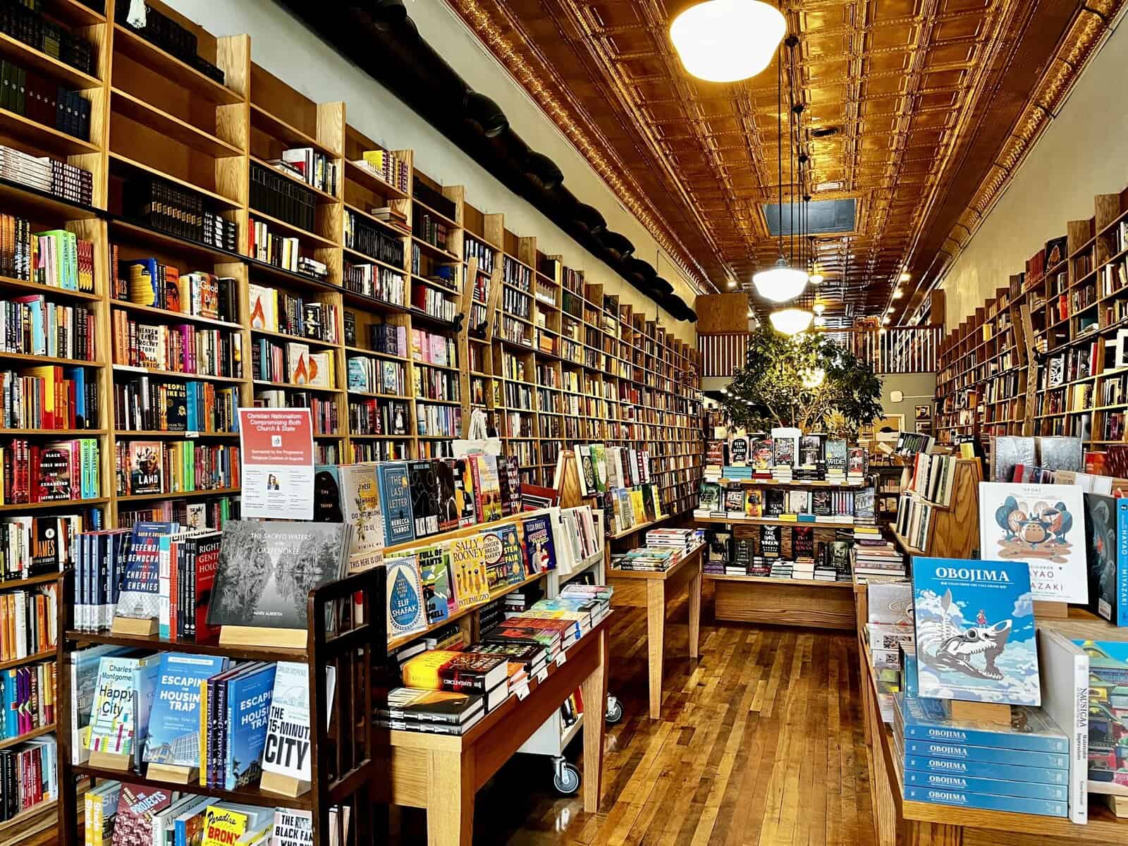 Interior of an independent bookstore featuring floor-to-ceiling wooden bookshelves lining both walls, decorative pressed tin ceiling, hardwood floors, and display tables with books and artwork in the foreground, with a small tree visible in the background of the long narrow space.