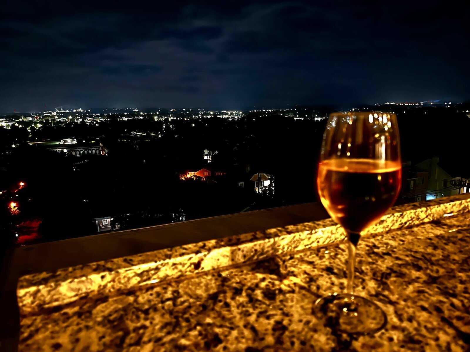 Wine glass sitting on a granite bar table in foreground with Augusta city lights twinkling across the dark landscape in the background at night, residential homes and buildings visible below.