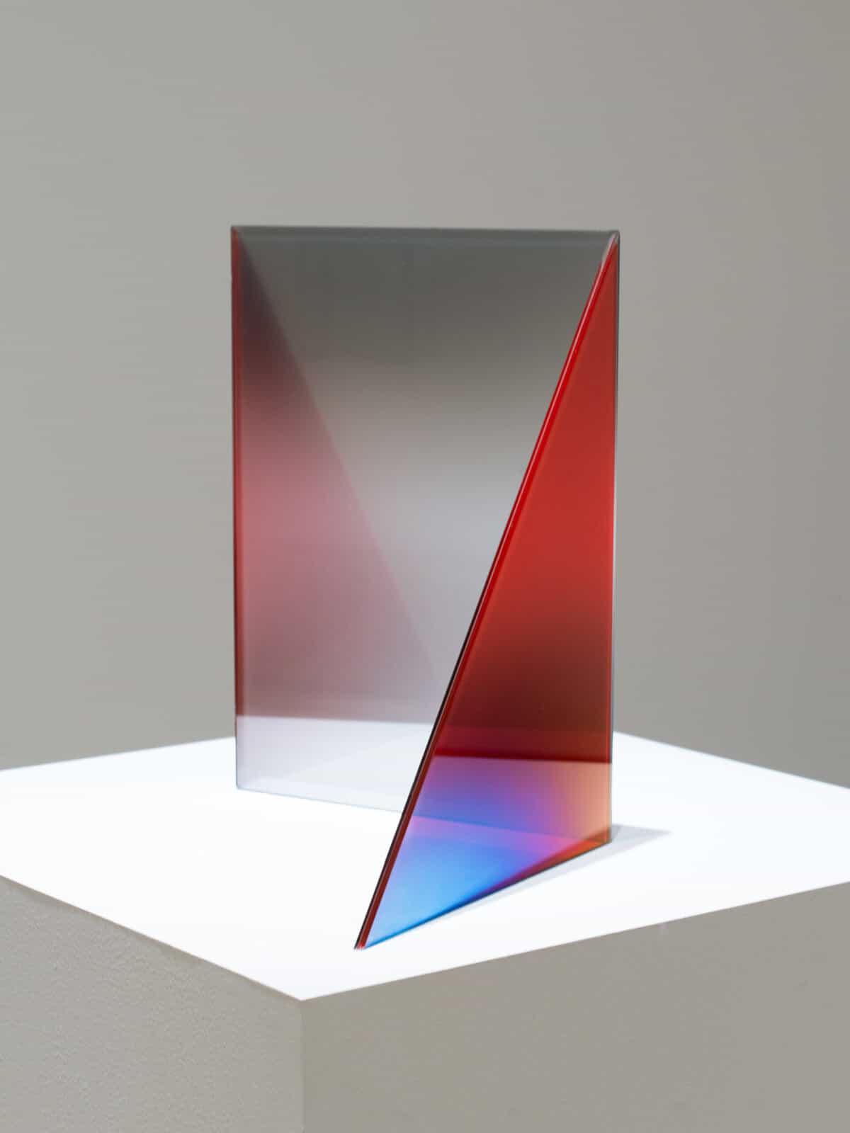 Minimalist glass sculpture by Larry Bell on a white pedestal: two thin, intersecting panels form a sharp triangular plane, with a gradient from gray to red and a vivid blue edge glowing where light passes through.