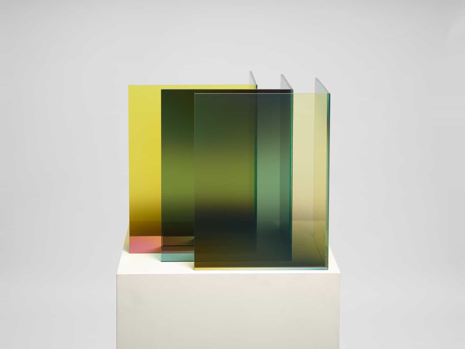  A minimalist sculpture by Larry Bell on a white pedestal: several parallel glass sheets with yellow-to-green gradients form a box-like stack, casting soft, layered reflections and shadows against a light gray backdrop. 
