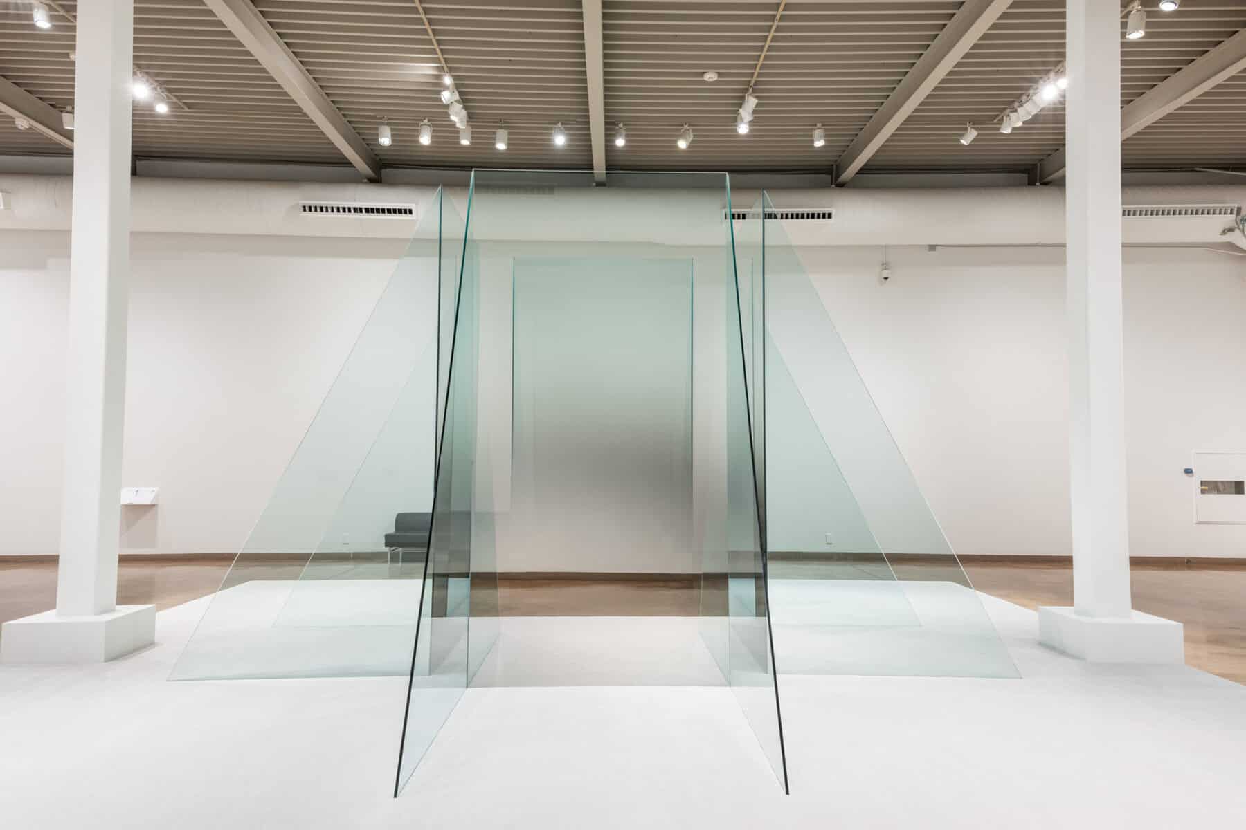 Large installation by Larry Bell consisting of tall, thin, transparent glass panels arranged in angled, trapezoidal planes that form a walk-through corridor; the pale green sheets reflect and overlap under bright gallery lights in a white-walled room with exposed ceiling beams.