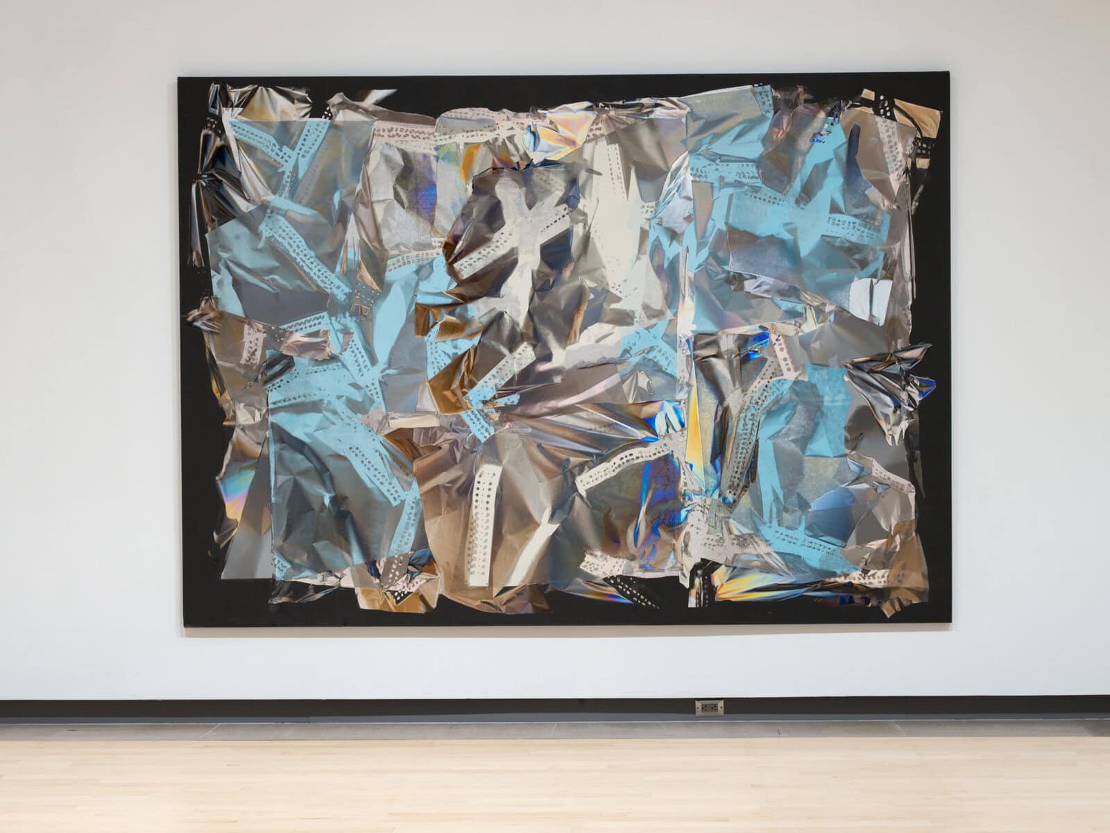 Large wall-mounted work by Larry Bell resembling a crumpled metallic collage: overlapping sheets of reflective silver film with patches of pale blue and iridescent highlights are arranged on a black rectangular backing, creating a shimmering, textured surface.