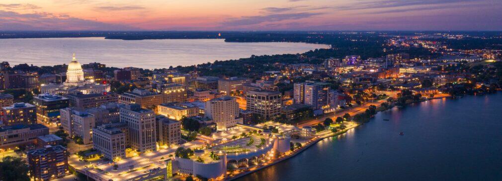 Aerial twilight view of downtown Madison showing the illuminated Wisconsin State Capitol building, surrounding city blocks lit up at dusk, and Lake Mendota in the background with the curved Monona Terrace convention center along the lakefront in purple and orange sunset light.