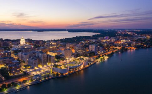 Aerial twilight view of downtown Madison showing the illuminated Wisconsin State Capitol building, surrounding city blocks lit up at dusk, and Lake Mendota in the background with the curved Monona Terrace convention center along the lakefront in purple and orange sunset light.