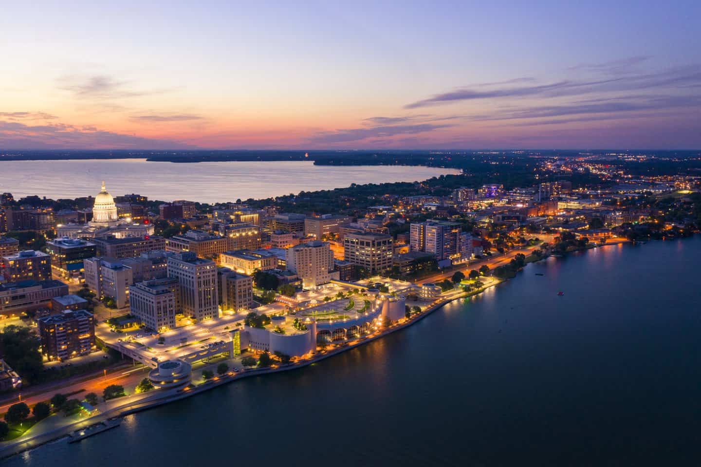 Aerial twilight view of downtown Madison showing the illuminated Wisconsin State Capitol building, surrounding city blocks lit up at dusk, and Lake Mendota in the background with the curved Monona Terrace convention center along the lakefront in purple and orange sunset light.