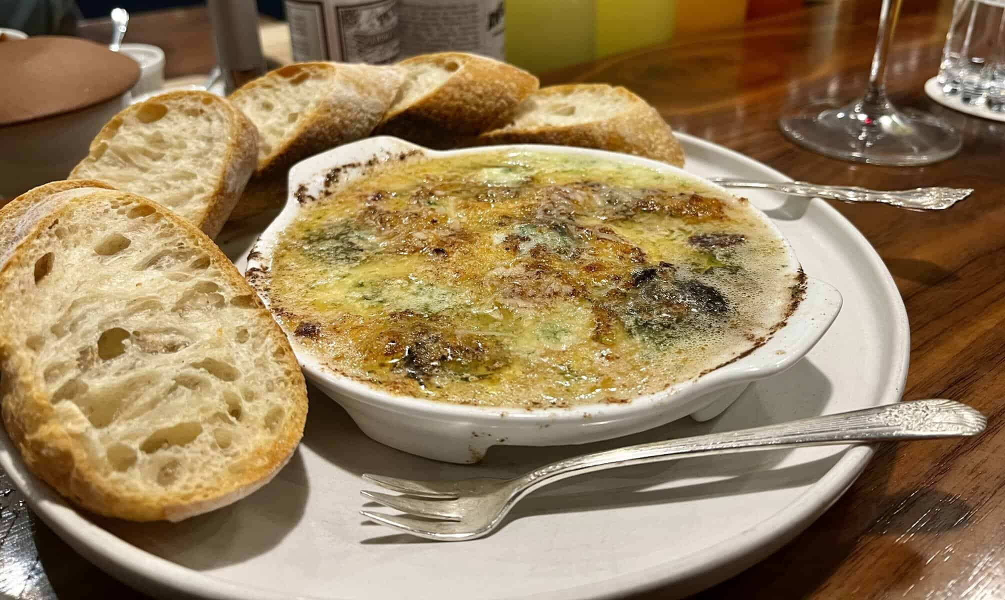 White oval dish containing escargots baked in garlic-parsley butter, served with sliced baguette arranged around the edge of the plate, photographed on a wooden table with glasses visible in the background.