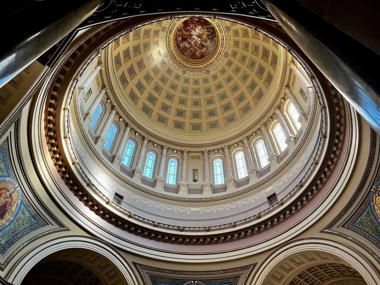 Upward view of the ornate dome interior of the Wisconsin State Capitol, featuring coffered ceiling panels, arched windows, decorative molding, and a painted oculus at the center surrounded by gold leaf detailing.