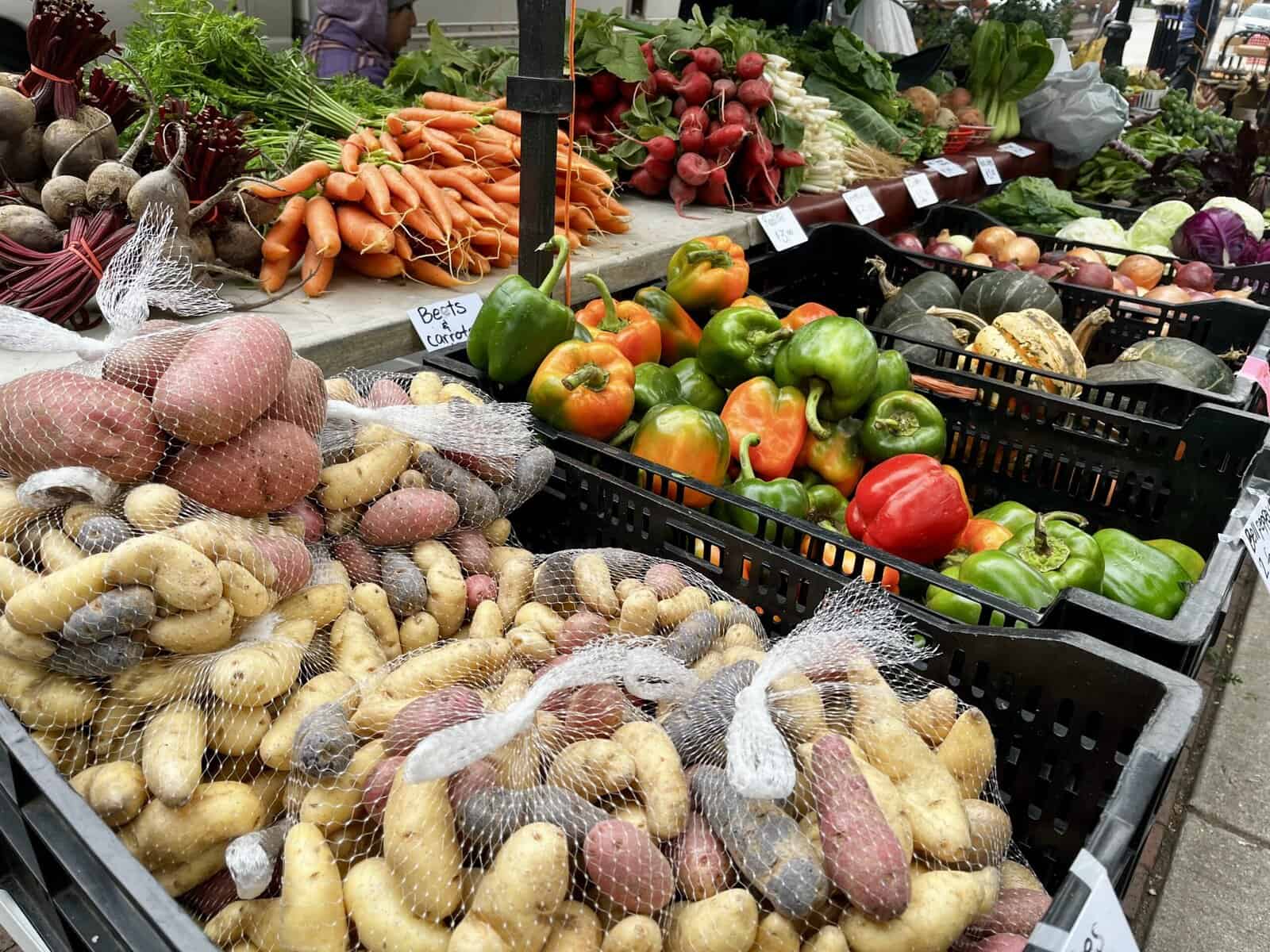 Colorful display of fresh produce at an outdoor farmers market including mesh bags of yellow and red potatoes in the foreground, bunches of carrots, beets, bell peppers in black crates, radishes, and various greens with shoppers browsing in the background.